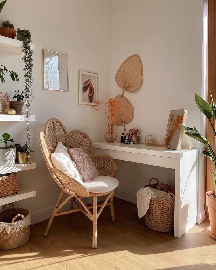 Decoração de verão de um canto aconchegante de uma sala. Há uma cadeira de vime com almofadas brancas e rosa claro, posicionada ao lado de uma mesa branca. Na parede acima da mesa estão pendurados dois quadros e um objeto decorativo em forma de folha. Uma variedade de plantas está presente na imagem; algumas estão em prateleiras montadas na parede à esquerda, outras sobre a mesa e no chão. A mesa branca contém objetos decorativos, incluindo um pequeno vaso com flores laranja secas. Um espelho retangular está apoiado contra a parede sobre a mesa. Dois cestos trançados estão no chão; um contém um cobertor dobrado. O ambiente é interno, possivelmente uma sala de estar, banhada por luz natural.

