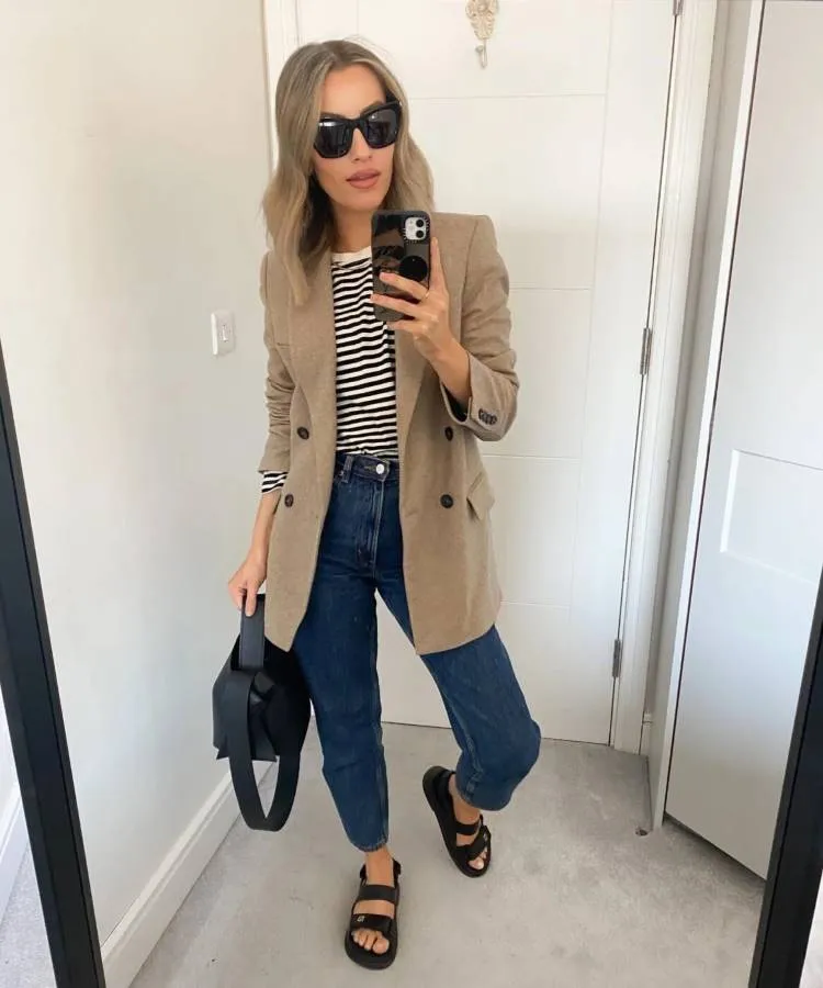 Uma mulher branca e loira posa em um espelho, ela usa um blazer bege, uma calça jeans e um sapato preto 