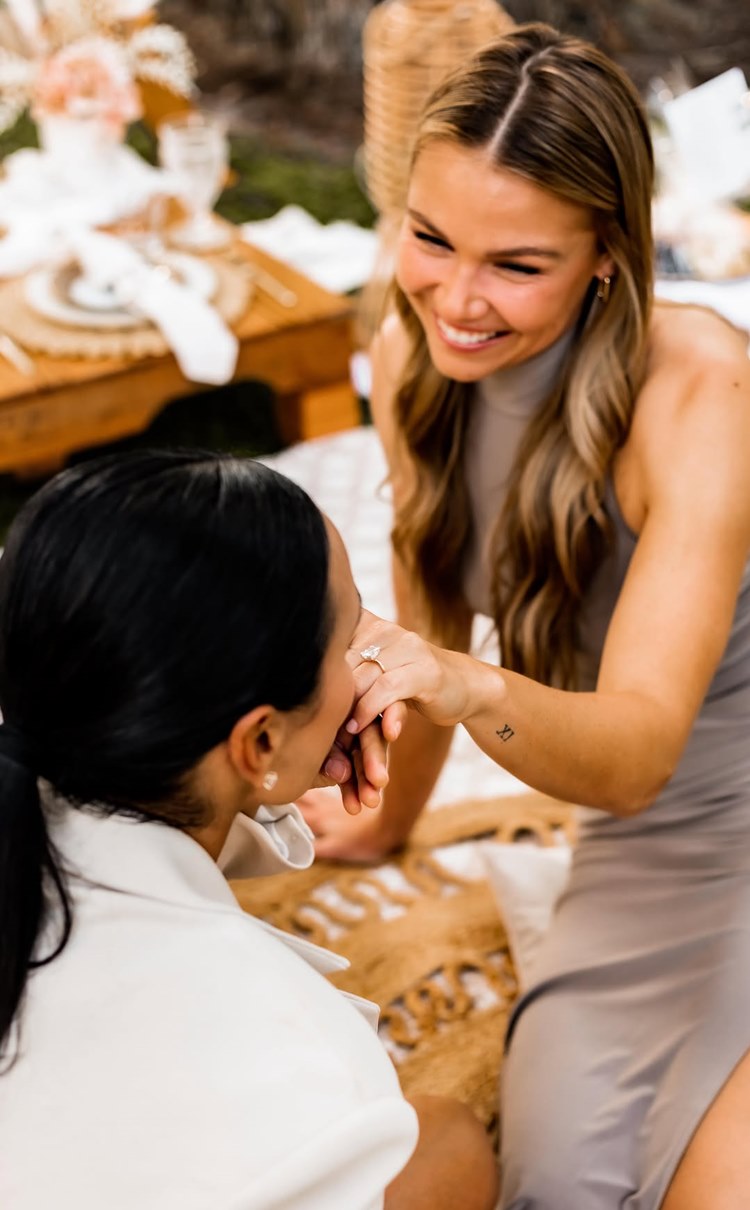 Duas mulheres, possivelmente amigas, estão juntas em um piquenique. Uma delas, sorrindo e com um anel de noivado no dedo, mostra-o à outra. A cena transmite felicidade e celebração entre as duas. Famosas que estão noivas