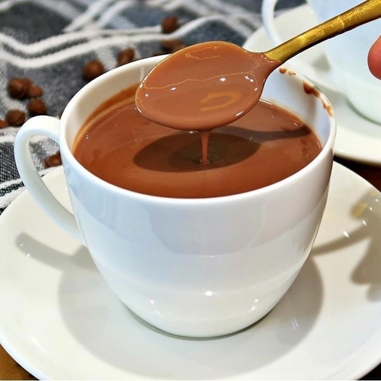 Foto de chocolate quente em xícara branca, com pires de mesma cor e colher dourada