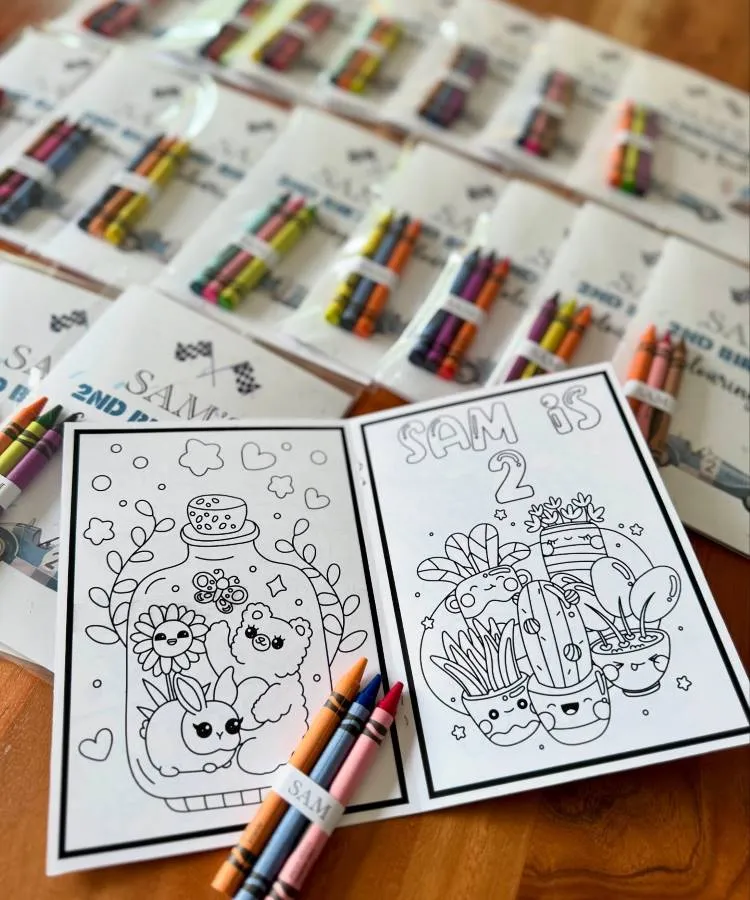 Livros de colorir personalizados com o nome "SAM" e a idade "2". Vários kits de giz de cera, cada um com quatro cores, estão dispostos ao redor, embalados e prontos para serem dados como lembrancinhas.

