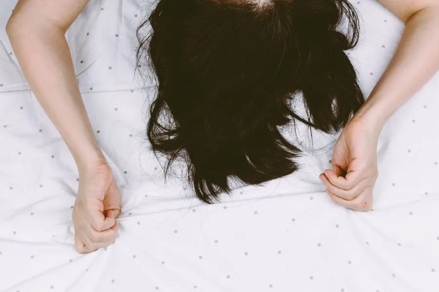 mulher deitada na cama com as duas mãos fechadas em punho agarrando o lençol branco