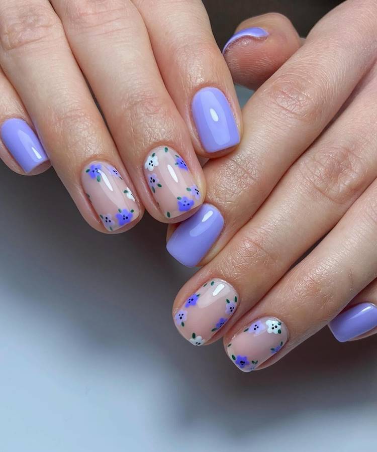 Unhas em esmalte roxo sólido alternadas com nude decorado com pequenas flores azuis e brancas, além de detalhes verdes. O design delicado e alegre cria um estilo primaveril elegante, destacando detalhes sutis contra o fundo claro e liso.