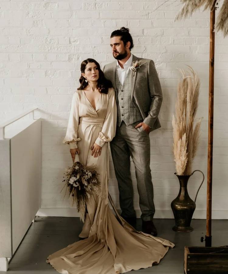 Um casal de noivos posa. A noiva usa um vestido de seda bege com decote V e cauda, segurando um buquê seco. O noivo veste um terno cinza xadrez. Ao lado, um vaso com plumas.







