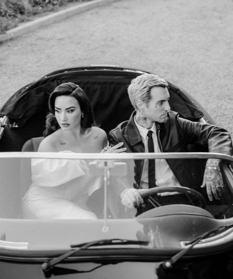 Em preto e branco, Demi Lovato e um homem estão em um carro conversível. Ela, de vestido de ombros caídos, olha para o lado, enquanto ele, de jaqueta de couro, está ao volante olhando para frente. A cena captura um momento clássico em um veículo antigo.