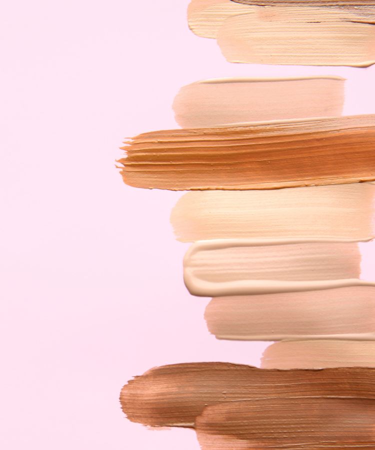 A imagem exibe várias amostras de base em diferentes tons, aplicadas em camadas horizontais sobre um fundo rosa claro. As cores variam de bege claro a marrom, com texturas cremosas e acabamentos lisos. Cada faixa de cor destaca a diversidade de tons disponíveis para diferentes tonalidades de pele.


