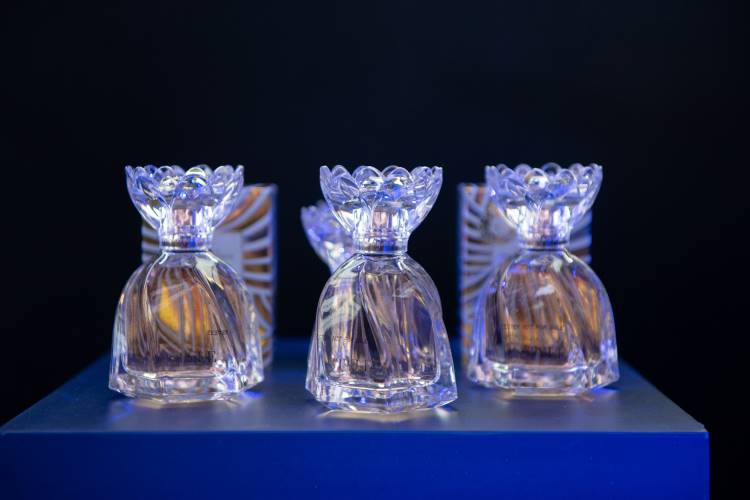 Frascos de perfume cristalinos, elegantemente iluminados, estão organizados sobre uma superfície azul. Cada frasco tem uma tampa decorativa e caixas ao fundo apresentam um padrão dourado e azul. O fundo escuro acentua o brilho e a sofisticação dos frascos.