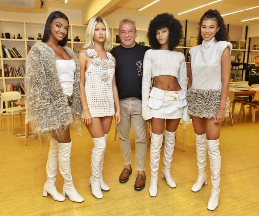 Claudio Silveira e algumas convidadas do coquetel estão em pé posando para foto em sala com estantes cheias de livros e mesas com cadeiras.