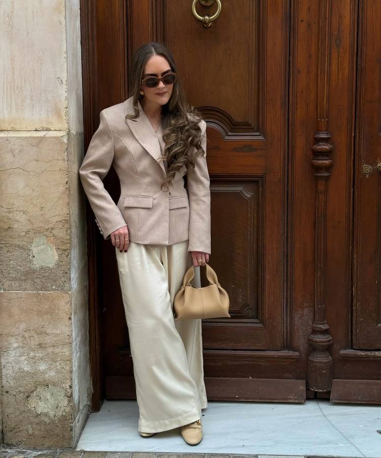 Mulher encostada em porta de madeira elegante, usa blazer nude ajustado, calça fluida off-white e sapatos nude. Segura bolsa bege minimalista e combina óculos escuros e cabelo solto ondulado. Estilo sofisticado perfeito para looks de trabalho com elegância discreta.





