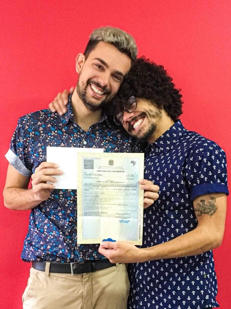 Vitor e Vinicius de camisa azul marinho abraçados segurando certidão de casamento 
