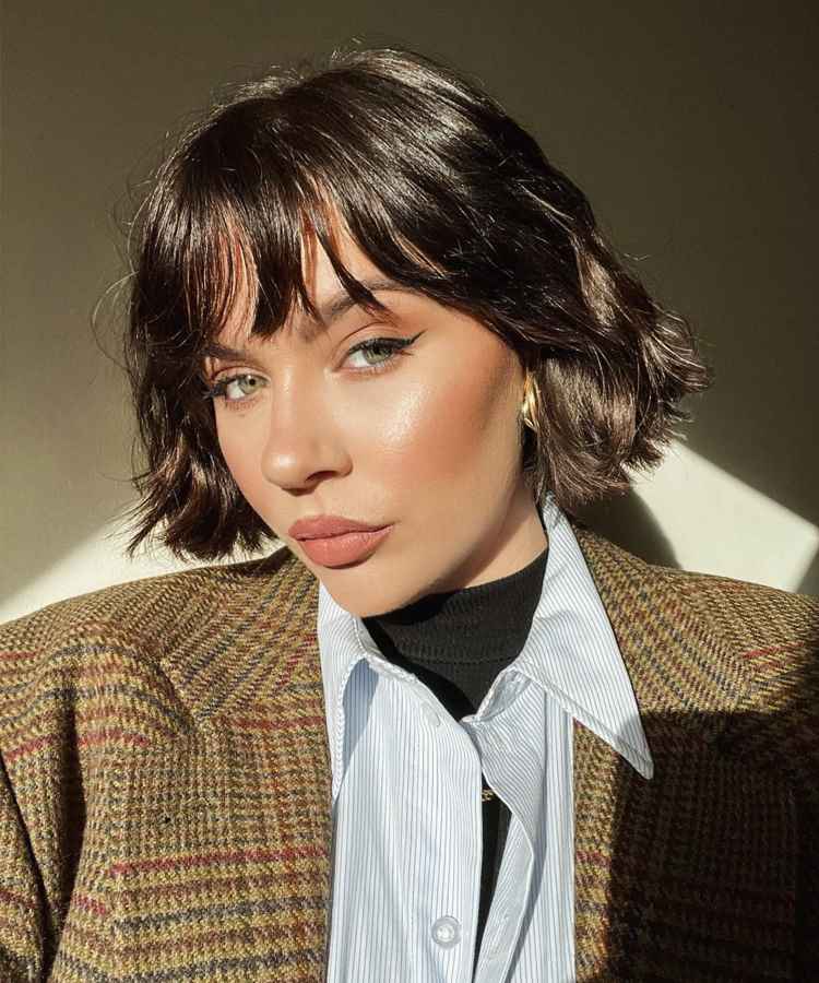 Uma mulher com cabelo castanho curto e ondulado, usando maquiagem discreta e delicada. A expressão é serena. Veste uma jaqueta xadrez marrom sobre uma camisa azul listrada e gola alta preta. A luz ilumina um lado do rosto.