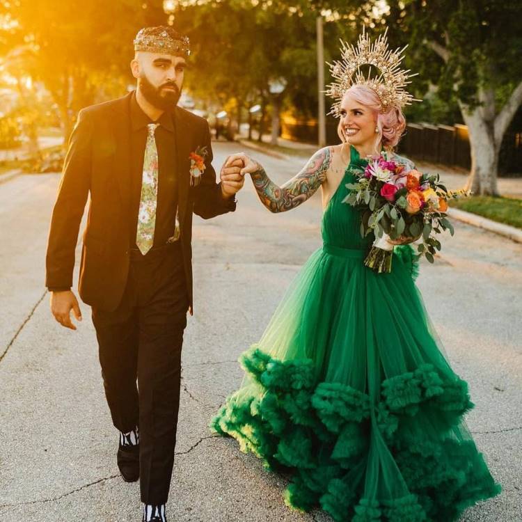 Uma noiva sorridente com cabelo rosa e coroa dourada, usando um vibrante vestido de tule verde com babados e mangas, de mãos dadas com o noivo de terno escuro e coroa, caminhando em uma rua ao pôr do sol.







