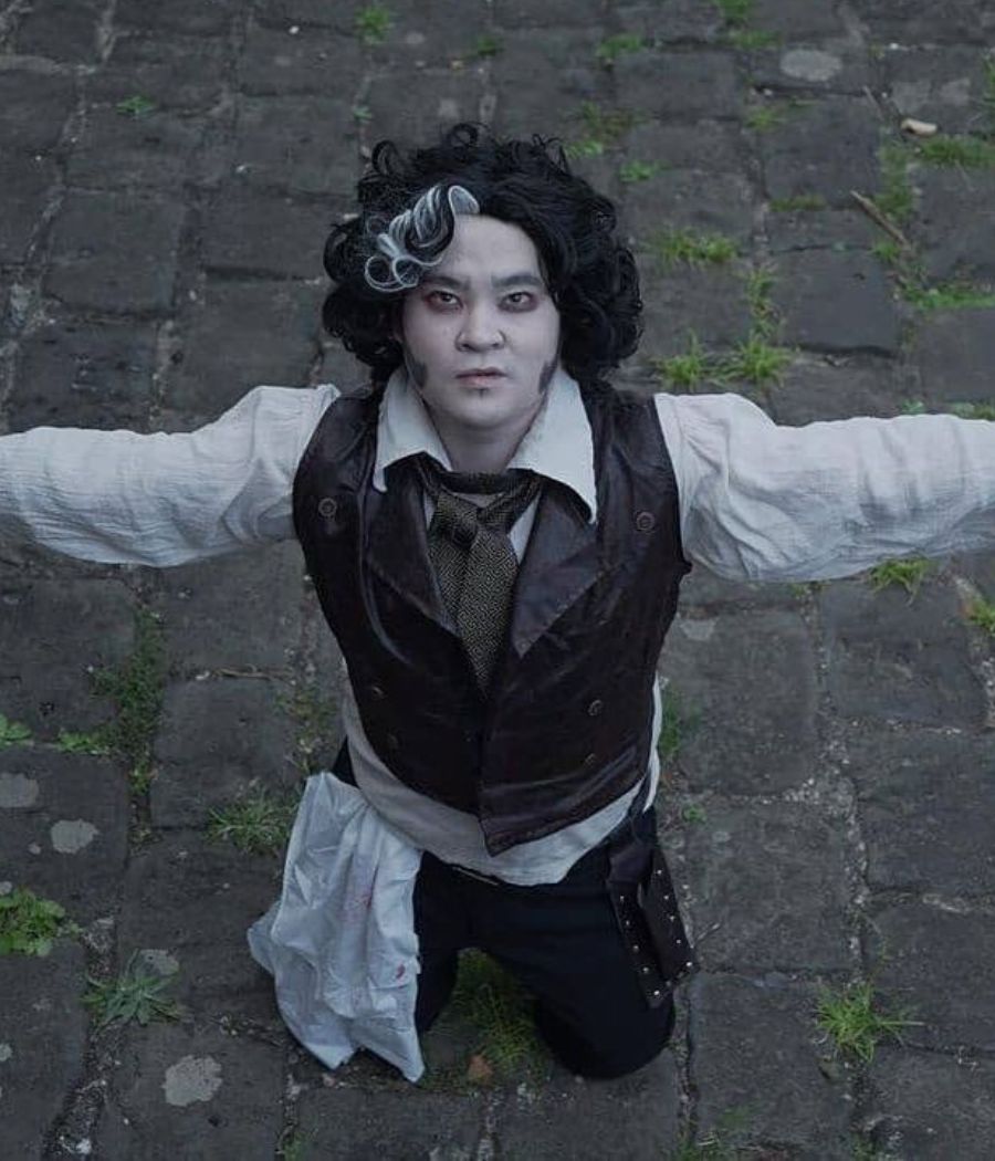 Homem asiático fantasiado de Sweeney Todd, com rosto pálido, cabelo preto e branco, camisa branca, colete marrom e calça 