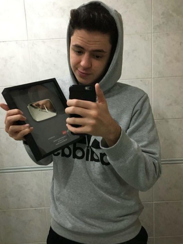 Enaldinho jovem tirando foto no espelho usando moletom cinza e segurando placa de 100 mil inscritos no YouTube