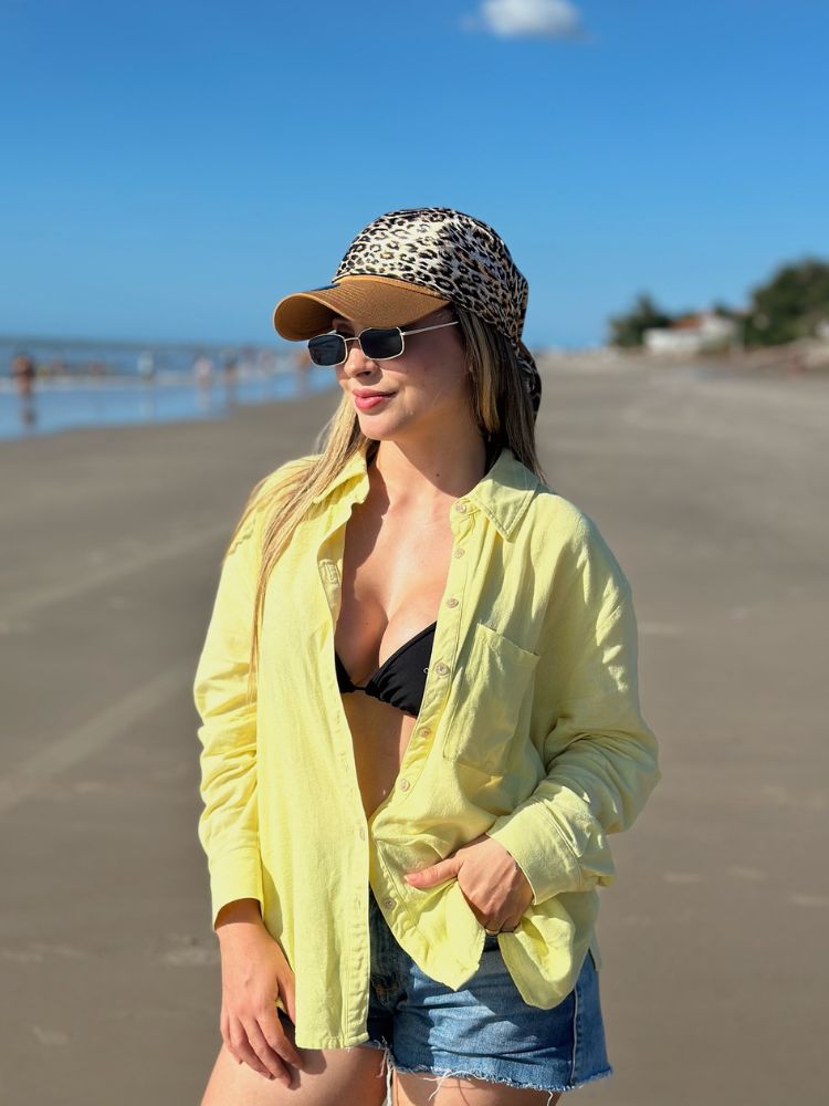 Mulher de pele clara na praia usando camisa amarela, biquini, boné marrom com lenço de animal print
