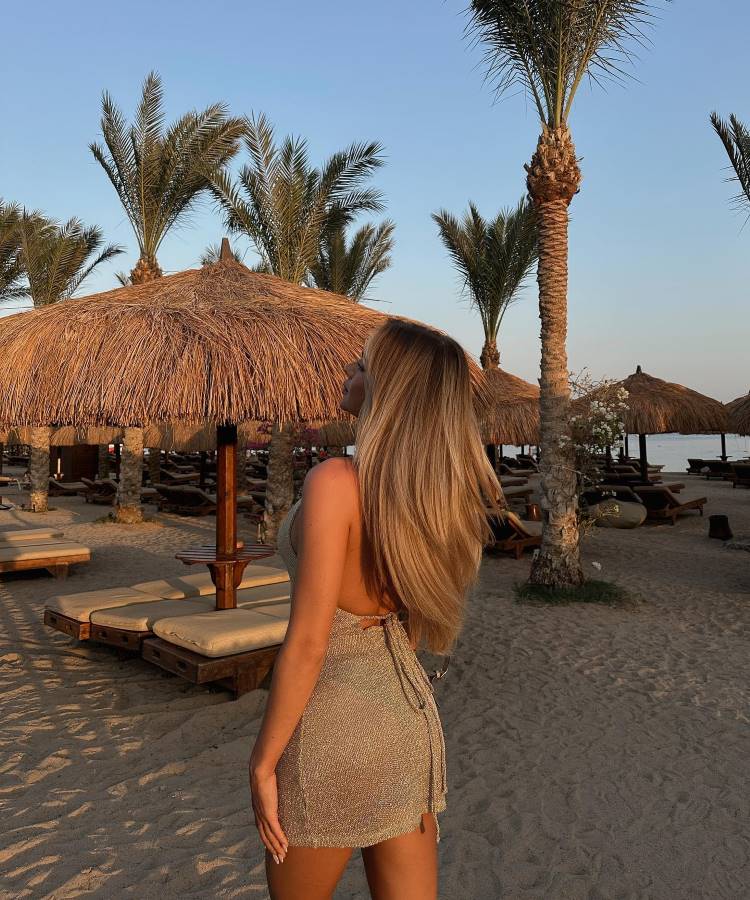 Uma mulher loira de costas, com cabelos longos e soltos, usa um vestido curto dourado. Ela está em uma praia com palmeiras e guarda-sóis de palha ao entardecer. A luz suave do sol cria uma atmosfera acolhedora, típica de fotos tumblr na praia.


