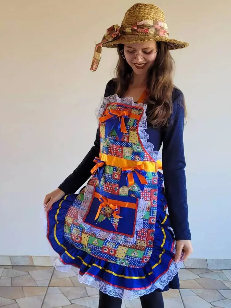 Mulher de pele clara usando blusa de manga longa azul, chapéu de palha r avental junino colorido, com bolsos, fitas e babados