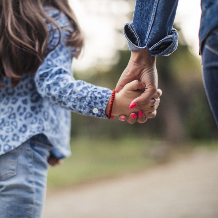 Uma mulher segura a mão de uma criança em um gesto de carinho e proteção. Ambas usam roupas casuais de jeans, destacando a simplicidade do momento em um parque com vegetação desfocada ao fundo, transmitindo conexão e ternura.