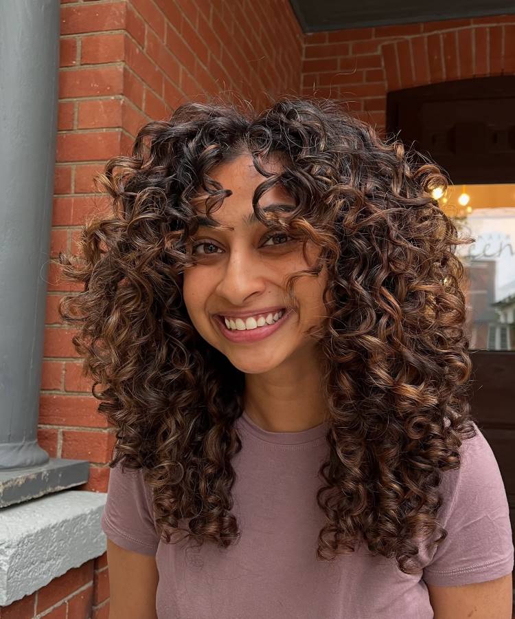 A imagem mostra uma mulher sorrindo, com longos cabelos cacheados e volumosos, em tons de castanho com reflexos dourados. Ela veste uma camiseta rosa discreta e está em um ambiente externo com uma parede de tijolos ao fundo.








