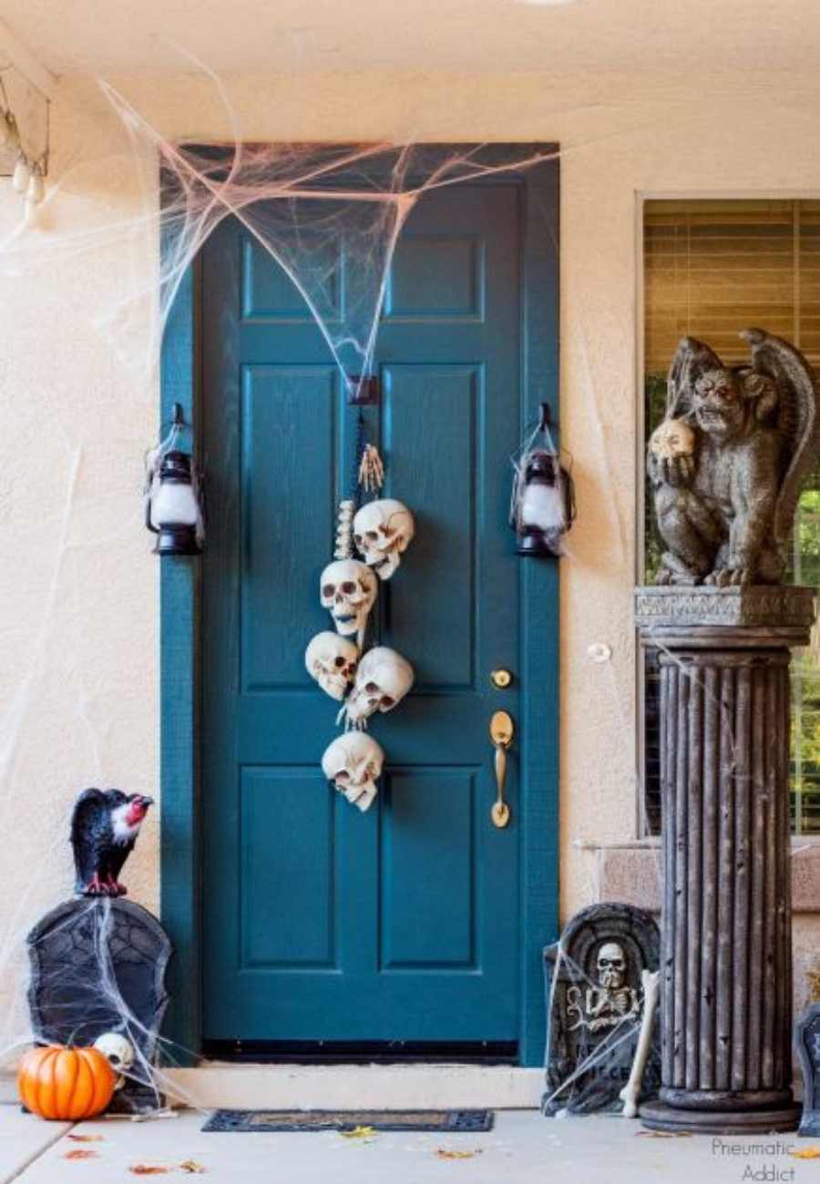 Entrada de casa com parede bege, porta azul com teias e caveiras, imitações de lápides e abóbora 