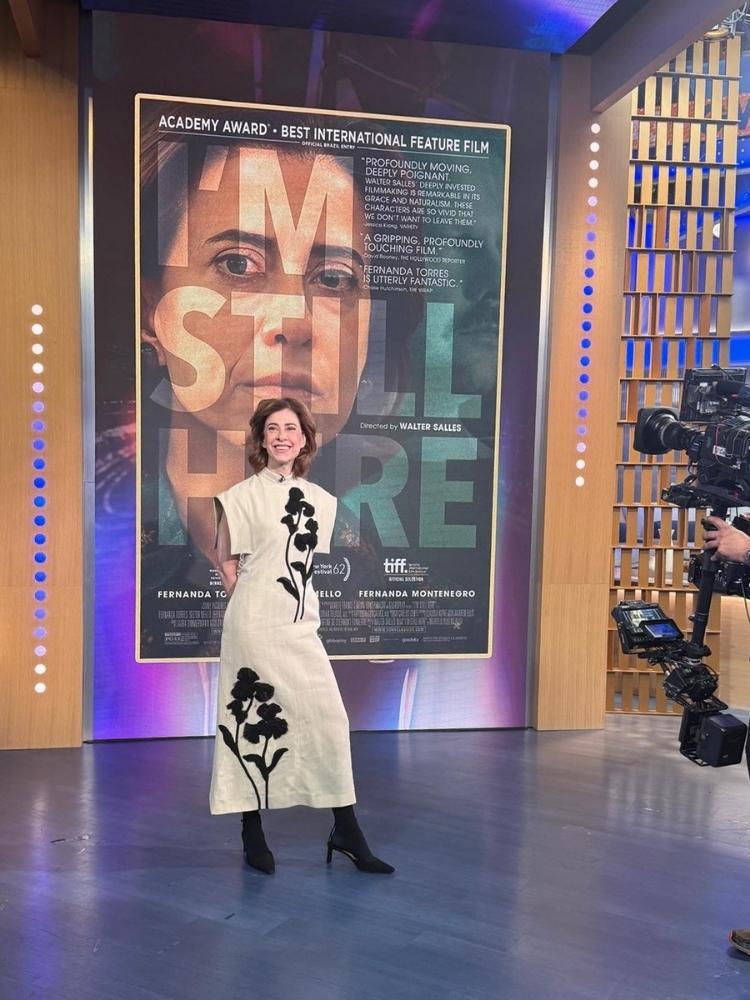 Fernanda Torres de vestido off white e preto em programa de auditório com cartaz de Ainda Estou Aqui ao fundo 
