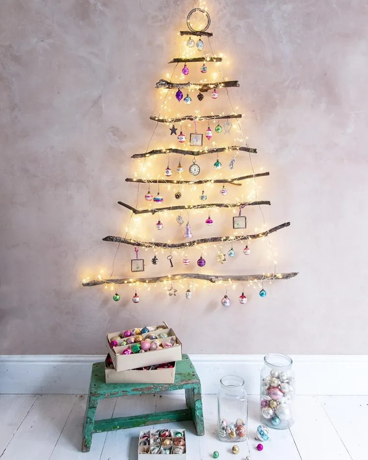 Parede cinza com árvore-de-natal de galhos com bolas natalinas e pisca-pisca. Abaixo, no chão, banco de madeira com caixas cheia de itens natalinos. Ao lado, potes de vidro com bolas de Natal.