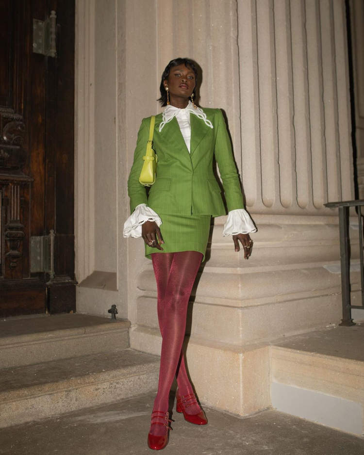 Mulher posando em frente uma coluna com roupa verde, meia calça vermelha e sapatos Mary Jane vermelhos de três tiras.