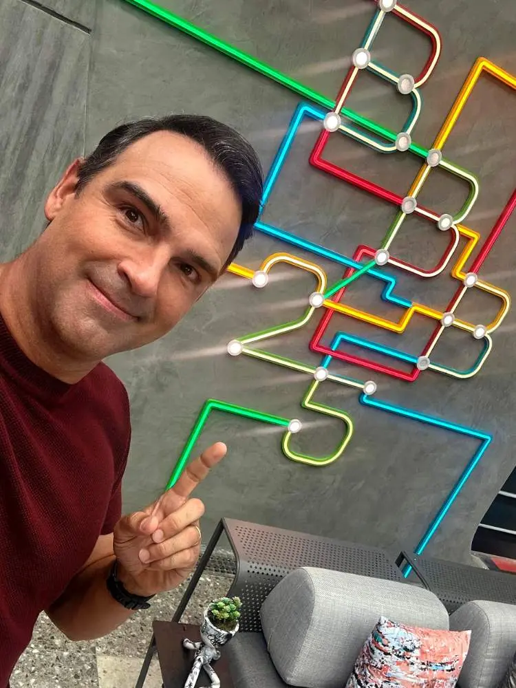 Tadeu de blusa vinho apontando para cenário com BBB 25 escrito por canos coloridos