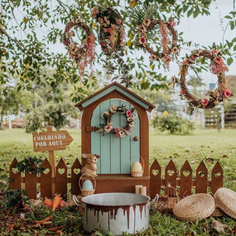 Quintal com grama e árvores com enfeite de Páscoa, com cerca de madeira, porta, guirlandas de Páscoa penduradas na árvore, coelhos e ovos de Páscoa