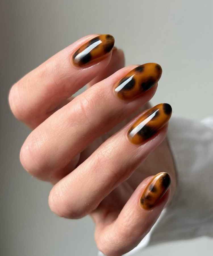 Unhas longas e ovais com design tartaruga em tons de marrom e preto. O acabamento é brilhante, destacando o padrão elegante. A mão está parcialmente fechada, e o fundo é neutro, o que ressalta ainda mais o estilo das unhas.