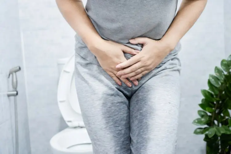 Pessoa de pele clara com calça e blusa cinzas, com as mãos no fim da barriga e banheiro ao fundo