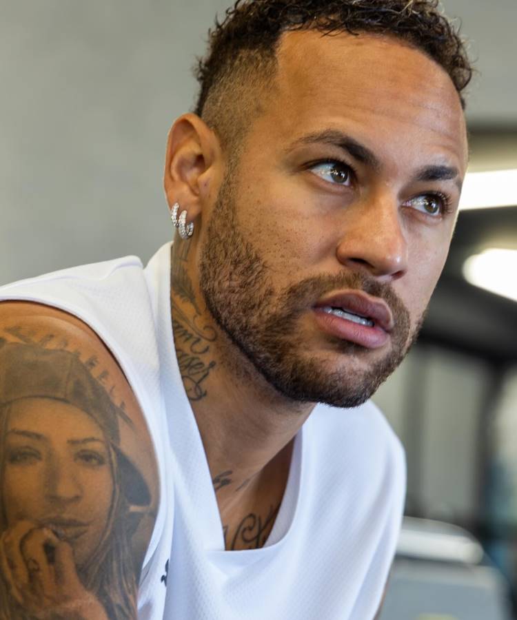 Neymar com cabelo curto e barba bem definida, usando camiseta branca. No braço, destaca-se uma tatuagem de uma mulher com chapéu olhando diretamente. Fundo desfocado sugere ambiente interno, possivelmente relacionado a treino ou academia.





