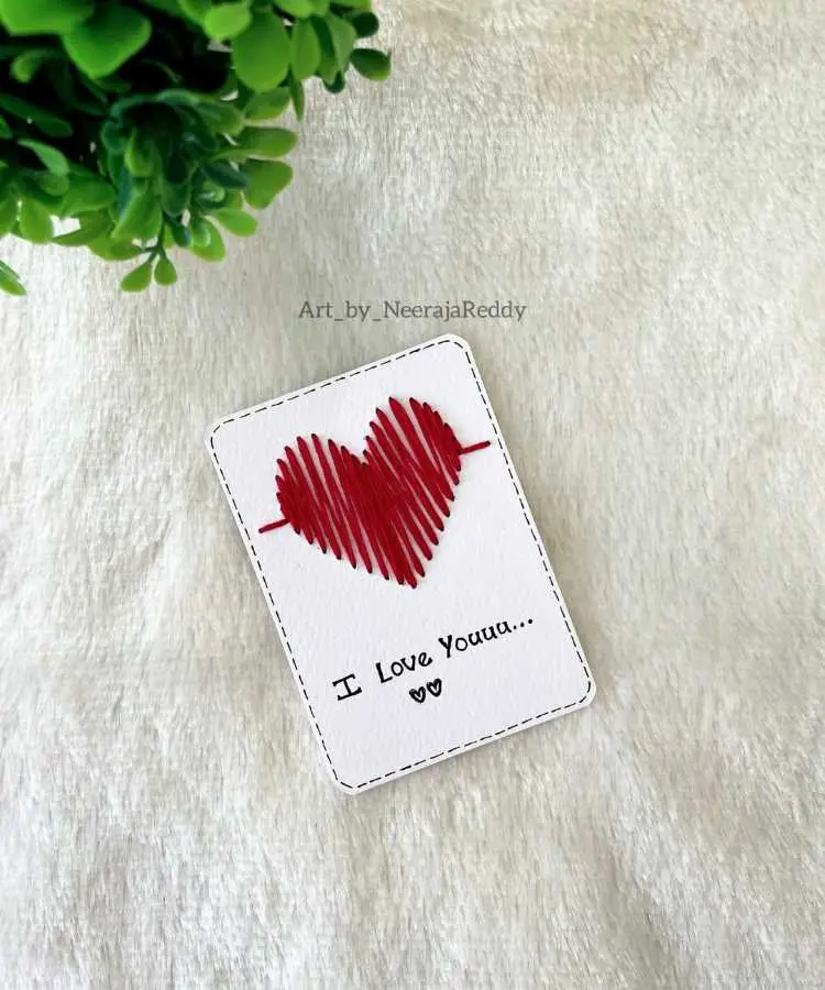 Cartão artesanal em papel branco com bordado de um coração vermelho e pequeno detalhe de linha preta nas bordas. Abaixo, a mensagem escrita à mão: "I Love You..." com dois corações desenhados. Fundo com textura de tecido claro.