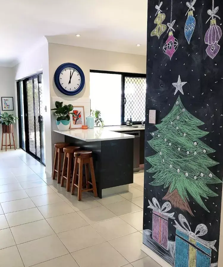 Cozinha moderna com piso claro e balcão preto com banquetas de madeira. Na parede, um relógio azul e quadro. Ao lado, uma lousa com desenho de árvore de Natal e presentes coloridos. Janelas grandes iluminam o ambiente, decorado com plantas.



