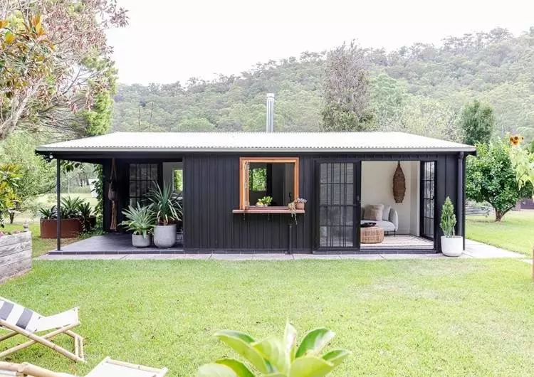 Imagem de fachada de casa simples feita em aço de cor preto. Ela está aberta, decorada com diversas plantas em vasos. Ao redor, gramado verde com cadeiras