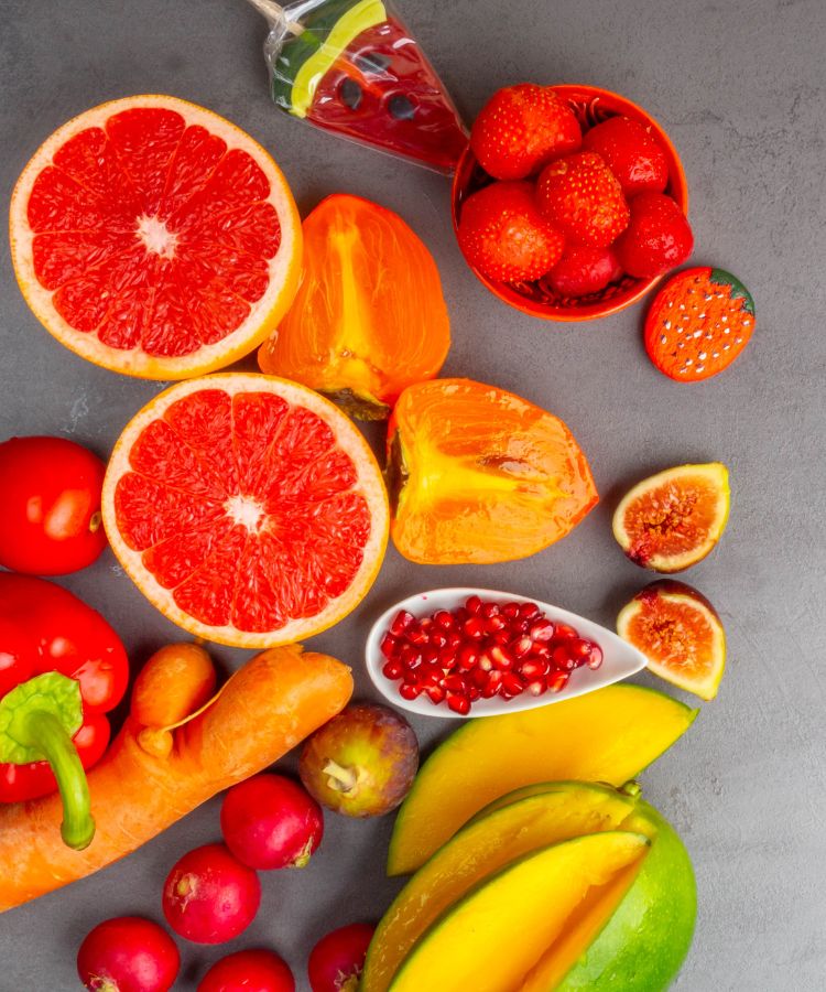 A imagem apresenta uma variedade de frutas e vegetais frescos e coloridos. Duas metades de grapefruit vermelha estão visíveis. Pedacinhos de caqui laranja brilhante estão próximos às grapefruits .Uma tigela pequena está cheia de morangos frescos e suculentos.Pimentões vermelhos inteiros adicionam outra camada de cor à composição. Uma cenoura laranja está presente na imagem. Grãos de romã são mostrados em um pote branco ovalado.





