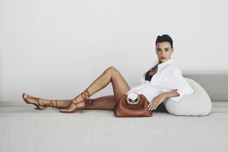Mulher morena deitada em um sofá, vestindo camisa branca e lingerie preta, com sandália marrom de tiras finas e bolsa da Arezzo, lança Habitat Natural, sobre o corpo, com as pernas cruzadas.