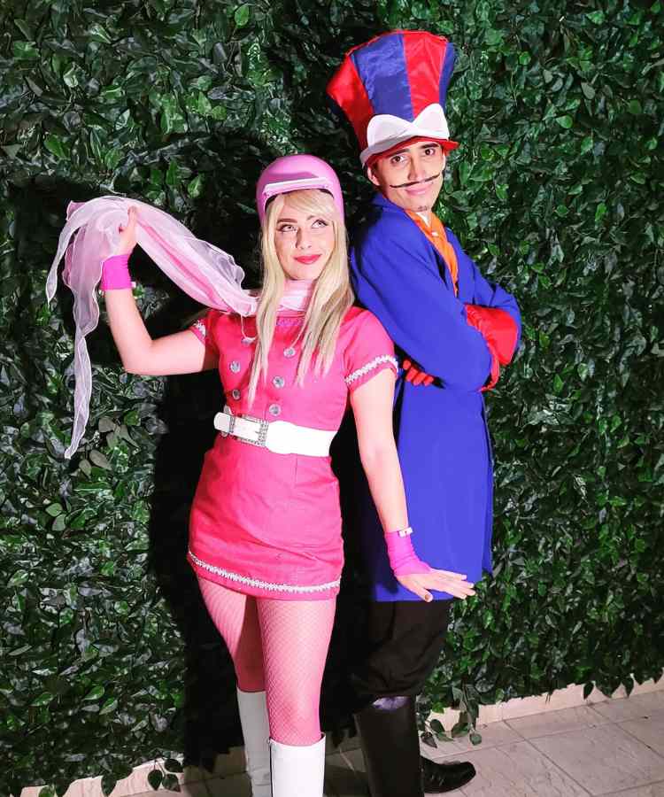 Traje de Dia das Bruxas para namorados: Penélope Charmosa e Dick Vigarista. Ela usa vestido rosa com cinto branco e capacete rosa, enquanto ele usa roupa roxa e chapéu roco e vermelho 
