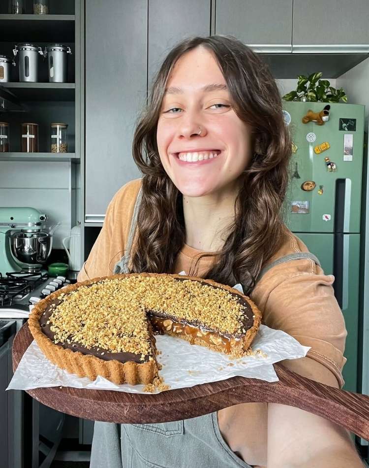 Foto de Laura Dicker usando blusa marrom com macacão jeans e segurando tábua de madeira com torta de chocolate, caramelo e amendoim