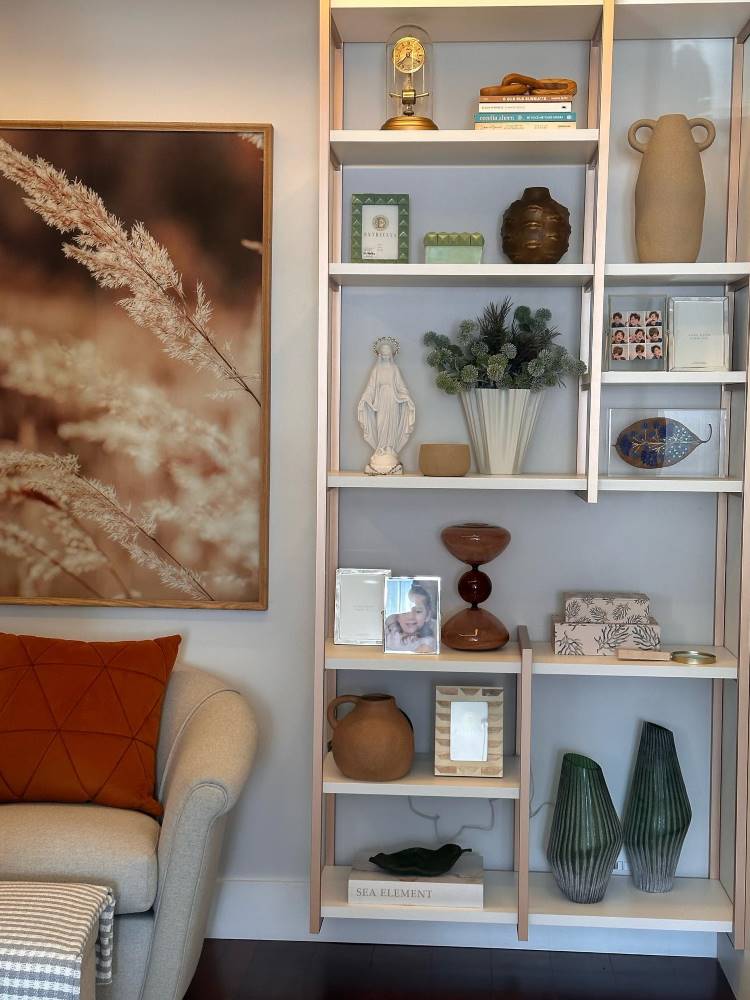 Estante vazada próxima ao sofá bege com almofada vinho. Nela, há diversos livros, vaso de planta, vasos, porta-retratos, quadros e diversos outros itens de decoração. 