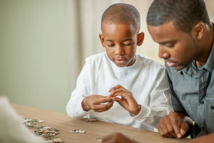 A imagem mostra um menino e um adulto montando um quebra-cabeça em uma mesa. O menino, concentrado, segura uma peça enquanto o adulto observa atentamente. Quebra-cabeças são excelentes presente para criança com autismo, ajudando no desenvolvimento cognitivo e na coordenação motora. O ambiente é calmo e propício para atividades educativas e interativas.