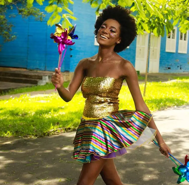 Mulher de pele negra usando cropped dourado, saia colorida e cataventos
