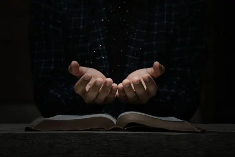 Duas mãos abertas, posicionadas acima de uma Bíblia aberta, parecem receber ou emanar pequenas partículas luminosas, criando uma sensação de espiritualidade. O fundo escuro e a luz focada intensificam a atmosfera de fé, mistério e contemplação.