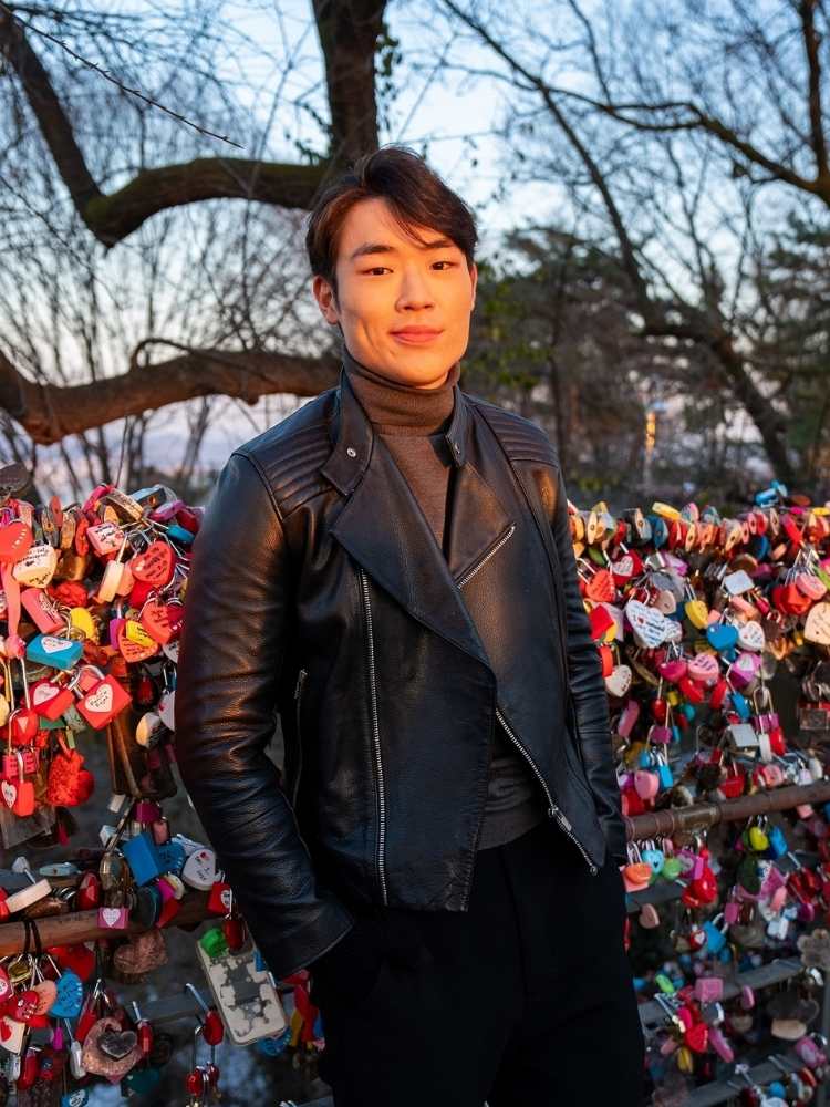 Arthur Paek usando blusa cinza de gola alta, jaqueta preta de couro em frente a ponte de cadeados na Coreia