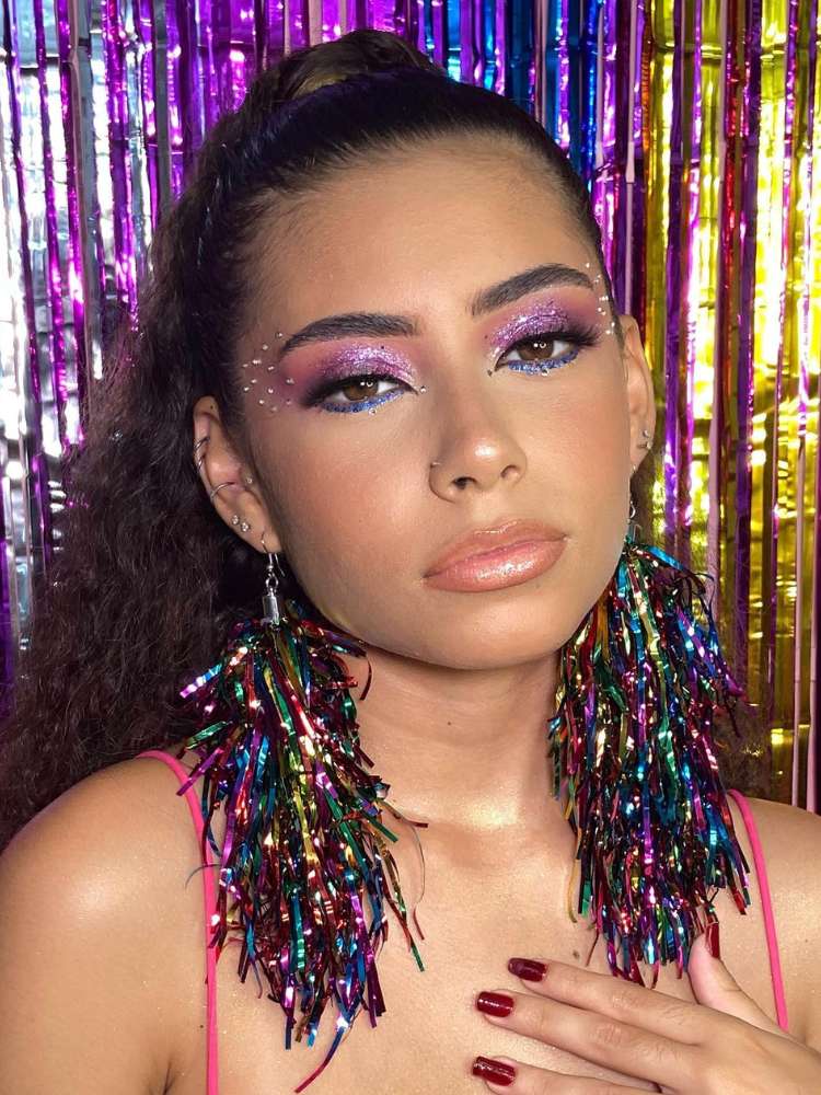 Mulher de pele clara com sombra roxa de glitter e brincos de fitas coloridas. 