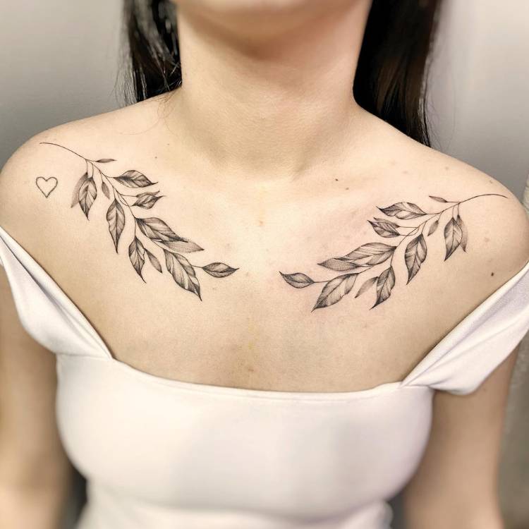 Mulher de pele clara e cabelo escuro usando regata bege e tendo dois ramos tatuados, um em cada ombro, além de um coração embaixo de um dos ramos