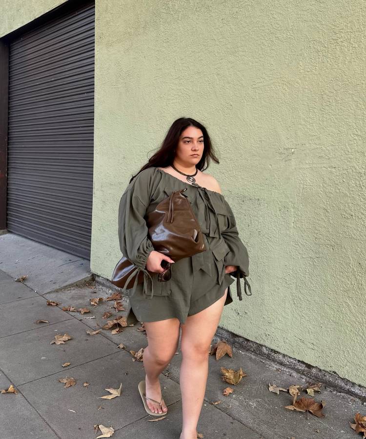 Uma mulher com cabelo escuro e comprido, vestindo um conjunto de short e blusa ombro a ombro de cor cáqui, caminha em uma calçada. Ela segura uma bolsa de couro marrom e usa chinelos, com o fundo desfocado.