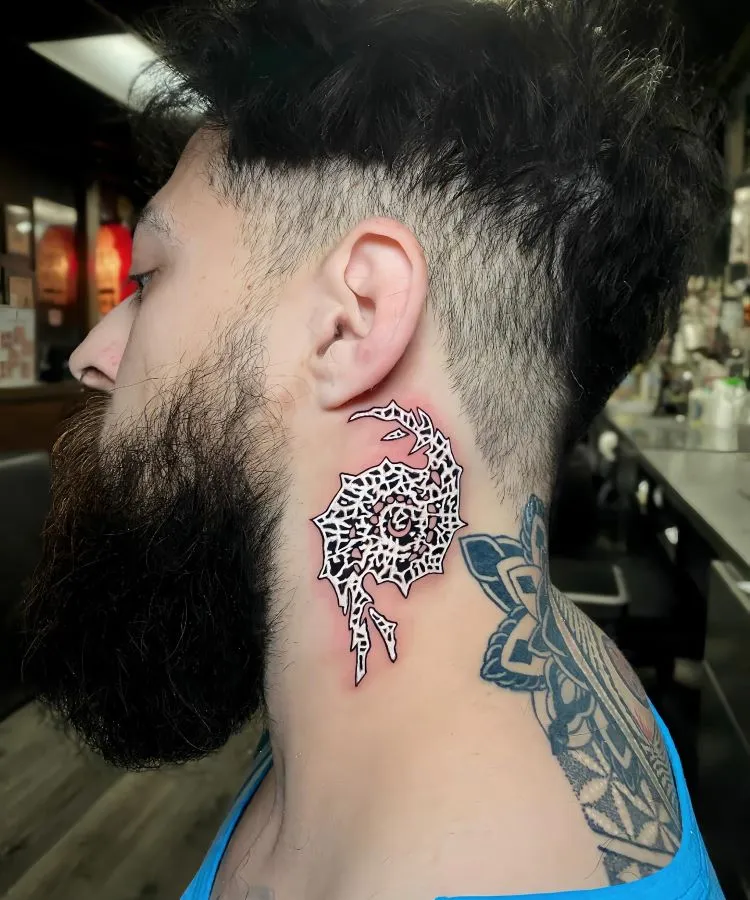 A imagem mostra uma pessoa de perfil com cabelo curto e barba. Ela tem uma tatuagem no pescoço de um design abstrato em preto e vermelho, parecendo um padrão tribal. A pessoa também tem uma tatuagem geométrica no ombro, visível pela camiseta azul cavada.


