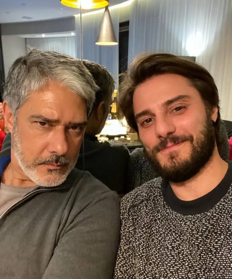 Dois homens posam para uma selfie. O mais velho, com cabelo grisalho e barba, usa uma jaqueta cinza e faz uma expressão séria. O mais novo, barbudo, sorri levemente para a câmera.