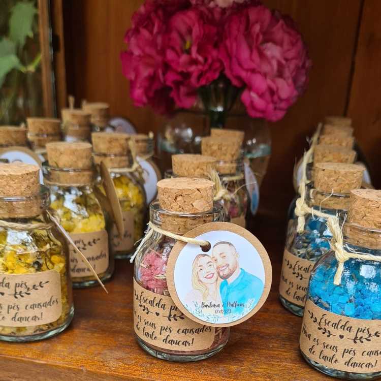 Pequenos frascos de vidro com tampas de cortiça, decorados com rótulos artesanais e cordões de juta, contêm sais coloridos. Cada pote traz uma tag ilustrada com casais personalizados. Ao fundo, flores rosas enfeitam o ambiente rústico e harmonioso.